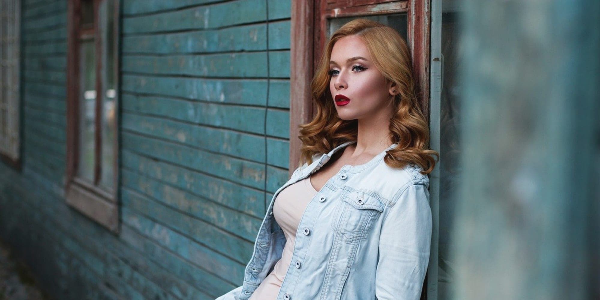 A woman in a denim jacket and jeans leaning against a painted timber wall